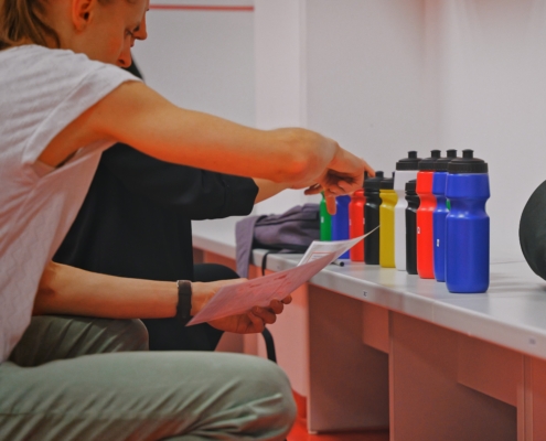 In einem Umkleideraum kniet eine Person vor einer Bank und zeigt auf eine Reihe bunter Trinkflaschen, die ordentlich nebeneinander aufgestellt sind. Die Flaschen haben verschiedene Farben, darunter Blau, Rot, Gelb, Grün und Schwarz. Die Person hält in der anderen Hand ein Blatt Papier, als würde sie etwas überprüfen. Auf der Bank liegen außerdem ein schwarzes Sporttaschen-ähnliches Objekt und einige Kleidungsstücke.