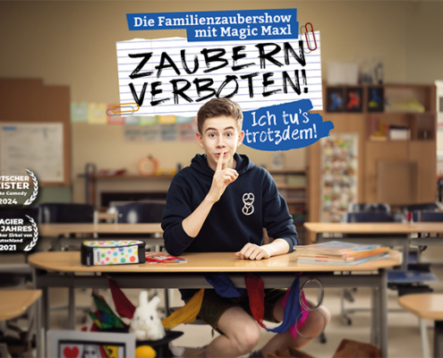 Der junge Zauberer Magic Maxl sitzt auf einer Schulbank im Klassenzimmer und hält sich den Zeigefinger vor den Mund. Pssst! Textblase: Zaubern verboten!