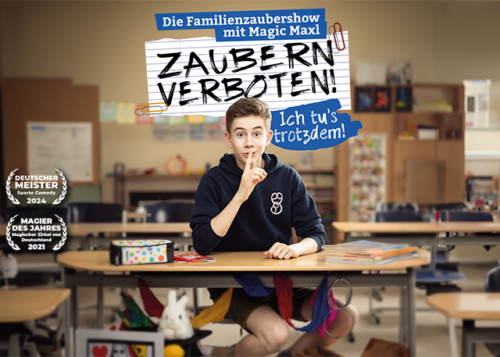 Der junge Zauberer Magic Maxl sitzt auf einer Schulbank im Klassenzimmer und hält sich den Zeigefinger vor den Mund. Pssst! Textblase: Zaubern verboten!